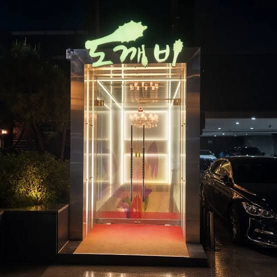 강남 도깨비 쩜오의 입구 전경, 밤에 초록색 도깨비 네온 간판 아래 흰 조명 테두리의 유리문 안으로 샹들리에와 붉은 바닥이 보임